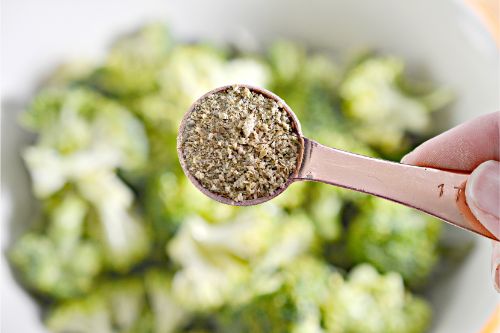 herbs in a tablespoon over broccoli in a mixing bowl 
