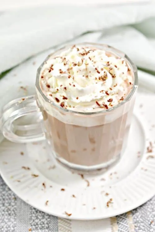 keto hot cocoa in a clear glass mug topped with whipped cream and chocolate powder on a white plate 