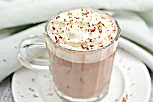 keto hot chocolate in a clear glass mug topped with whipped cream and chocolate powder on a white plate 