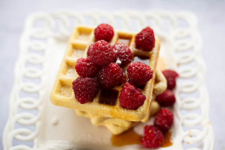 stack of keto waffles covered in raspberries and syrup sitting on a white lacy plate