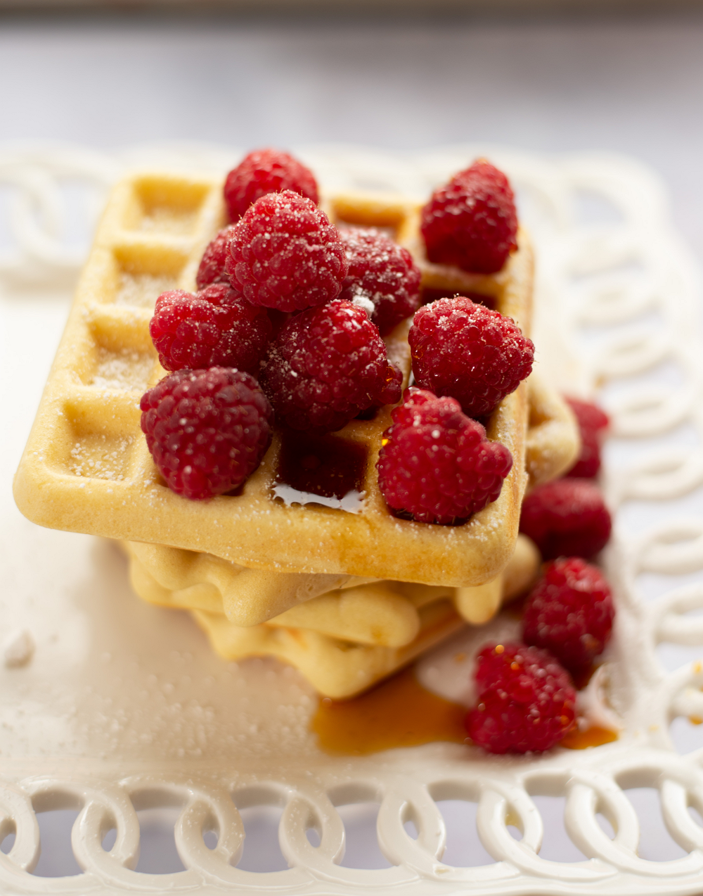stack of gluten-free waffles covered in raspberries and syrup sitting on a white lacy plate