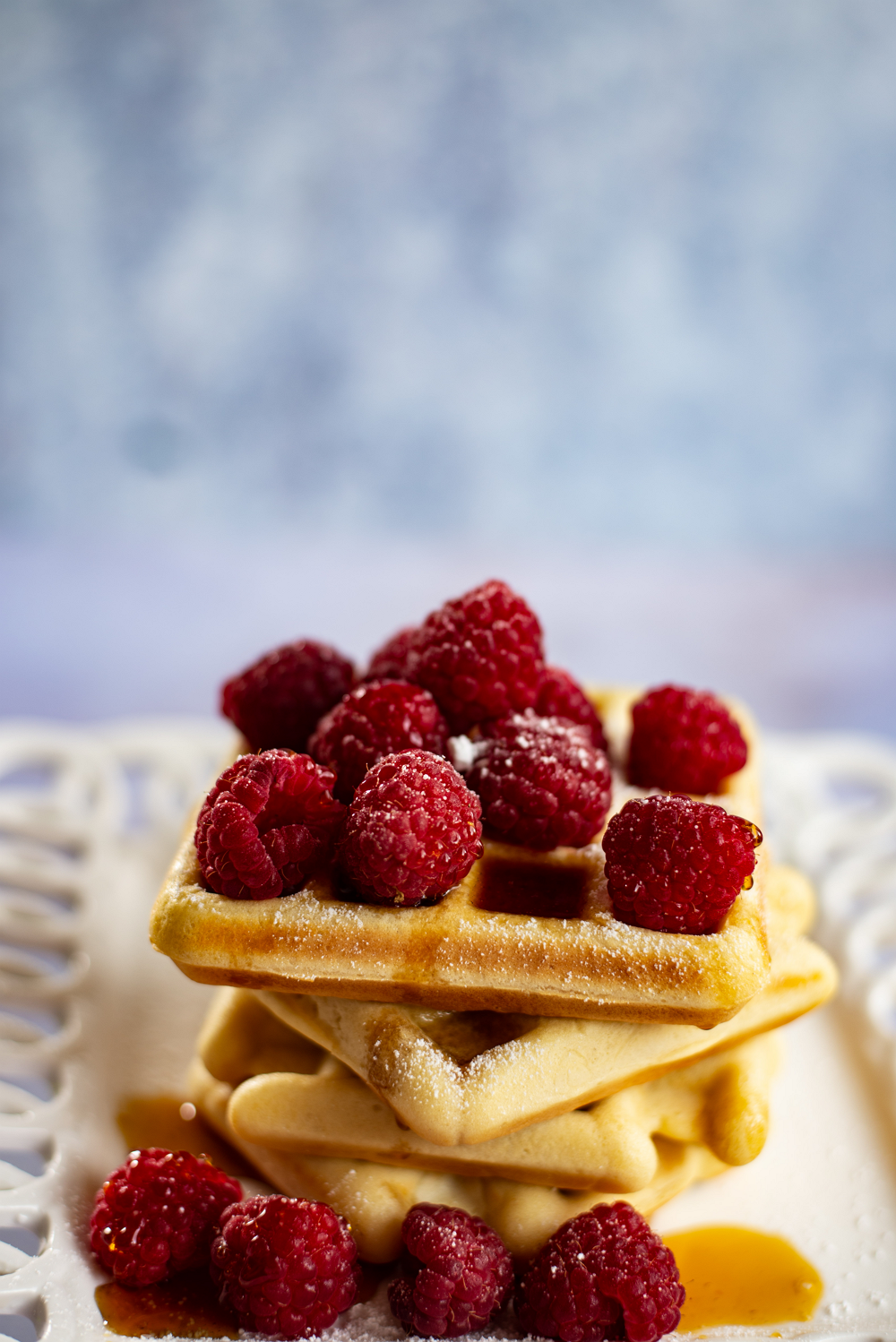 close up of stack of keto waffles covered in raspberries and syrup sitting on a white lacy plate