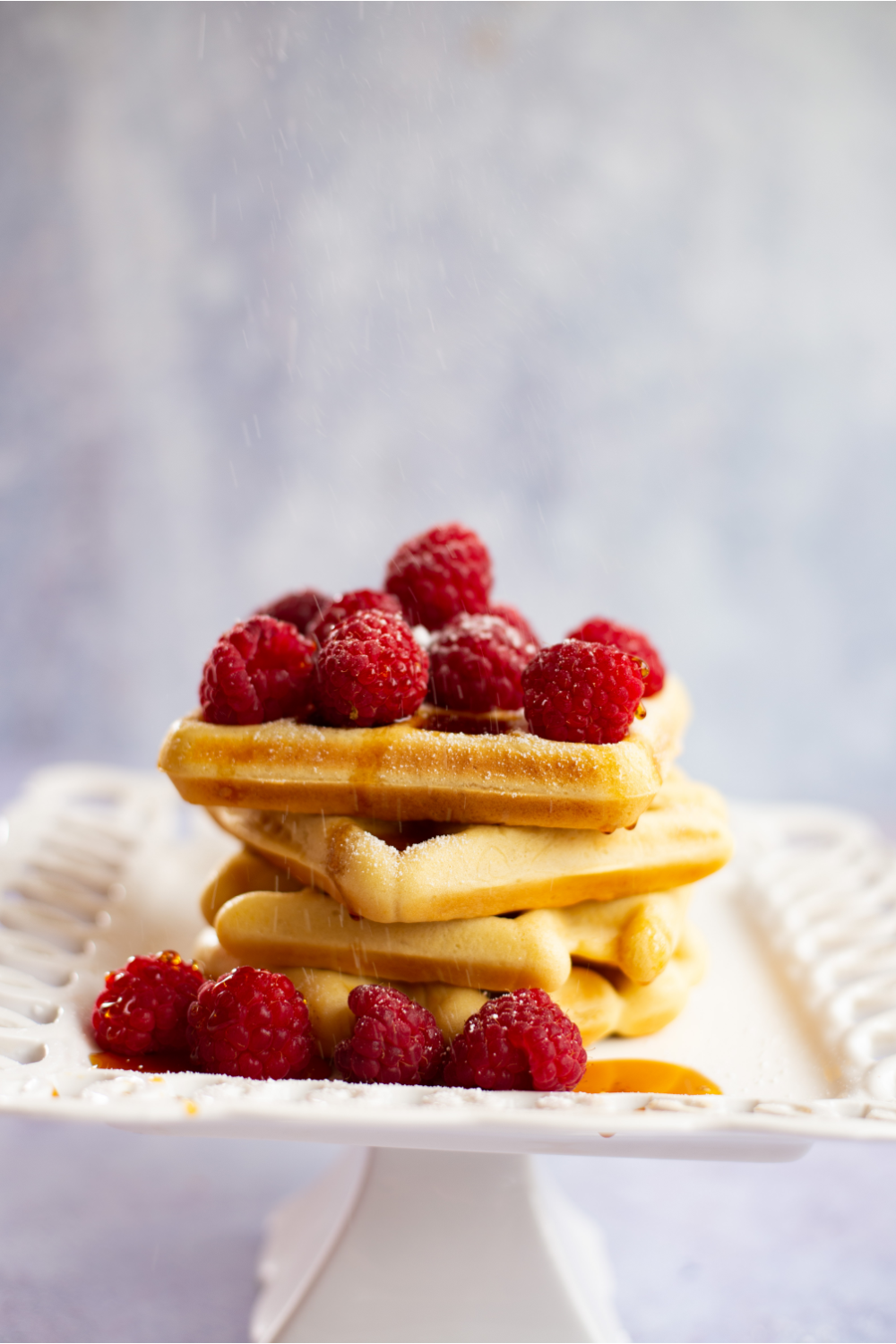 slanted shot waffles covered in raspberries and syrup sitting on a white lacy plate