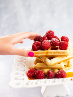 ladies hand with pink nail polish grabbing a waffle off a stack of waffles covered in raspberries and syrup sitting on a white lacy plate