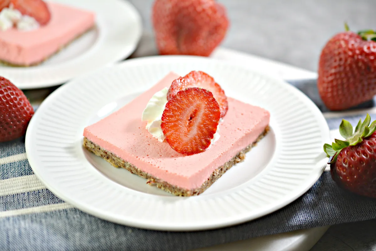 strawberry cheesecake bar cut and sitting on a white plate topped with a sliced strawberry