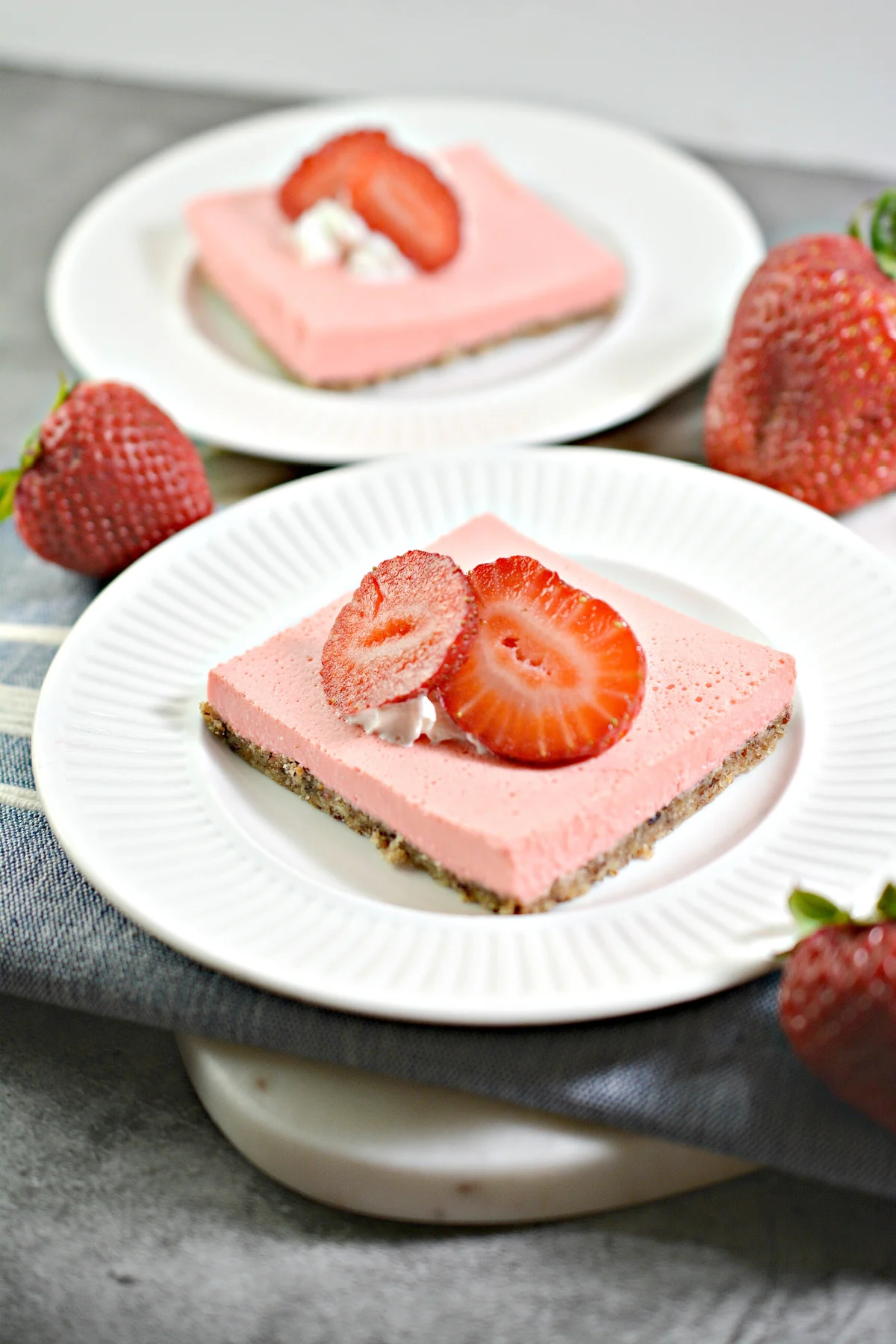 strawberry cheesecake bar cut and sitting on a white plate topped with a sliced strawberry with another plate in the background