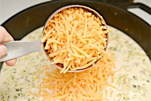 shredded cheese being poured into a pot of veggie and cheddar soup 