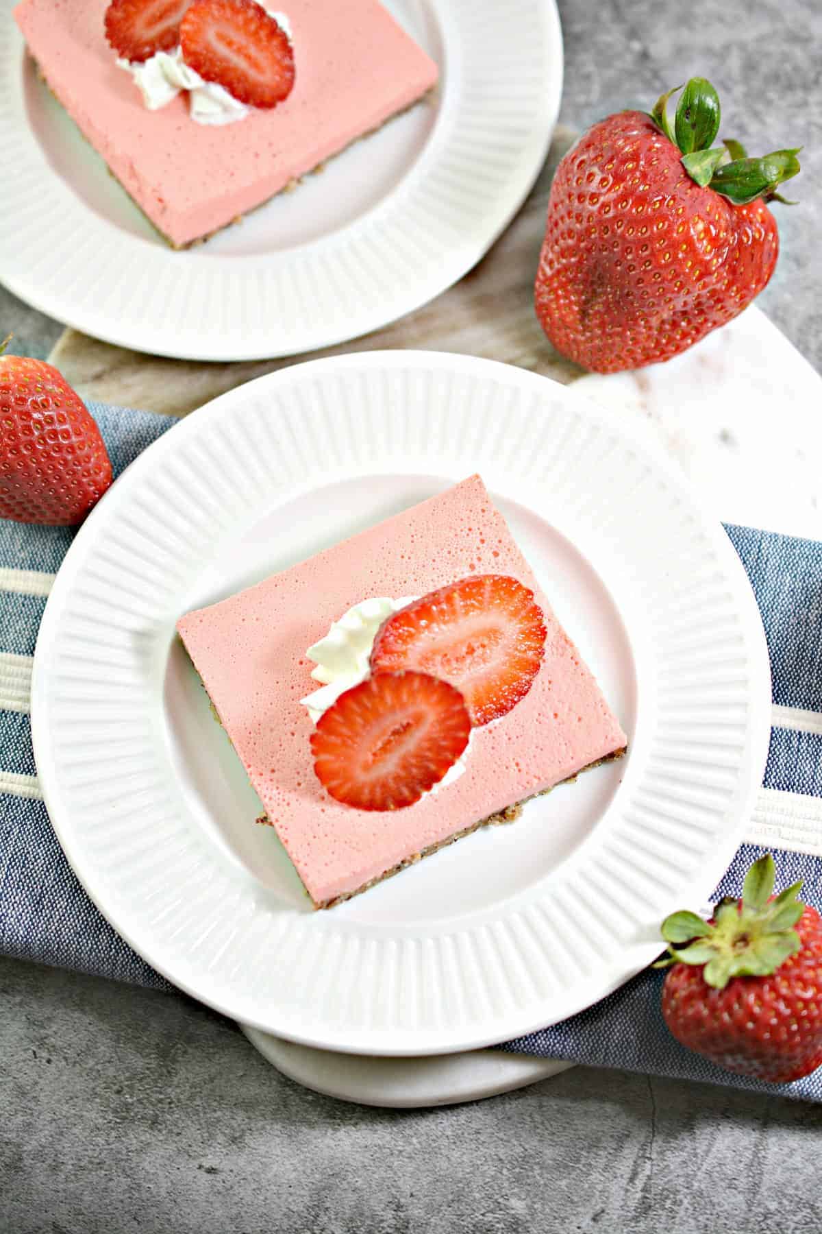 Two square slices of pink strawberry cheesecake bars on white plates, each topped with whipped cream and fresh sliced strawberries, with whole strawberries placed around the plates.